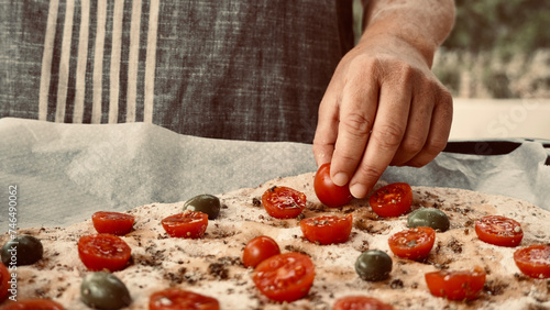 Woman preparing traditional Italian focaccia puts cherry tomatoes on top of the dough. Cooking outside in the garden with fresh ingredients.