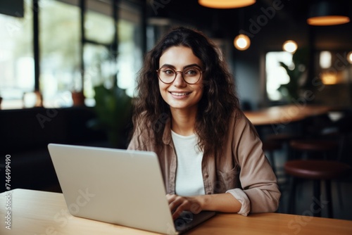 Cheerful woman in casual clothes and glasses sitting at table with laptop and looking at camera while working