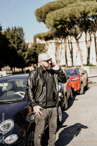 Happy smiling bearded man standing near a car at Rome,Italy.Man holding a mobile phone standing outdoors next to his car texting ,sending parking sms