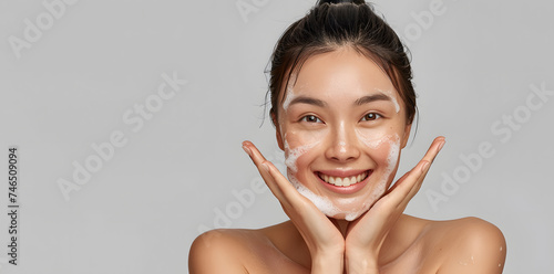 Close-up portrait of smiling  asian woman washing face with soap foam, isolated on light grey background