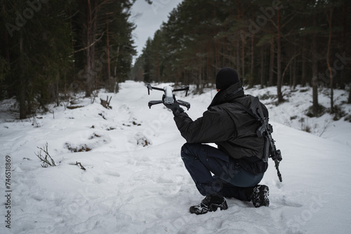 A military man with a rifle behind his back launches a drone in the forest in winter.