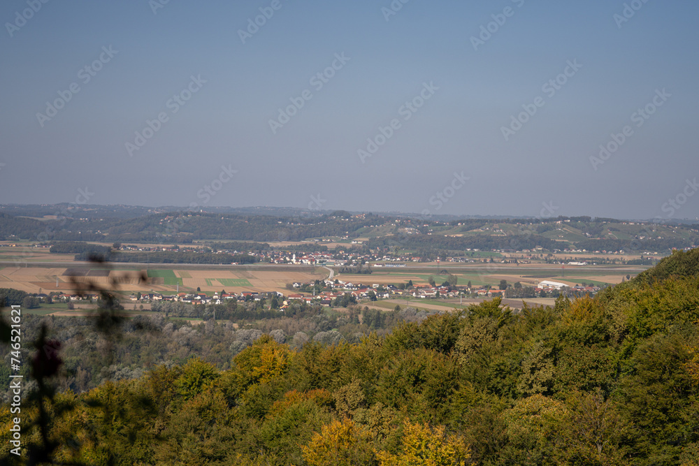 rural landscape unfolds from a high vantage point, with a patchwork of agricultural fields, a small village, and distant rolling hills under a hazy blue sky