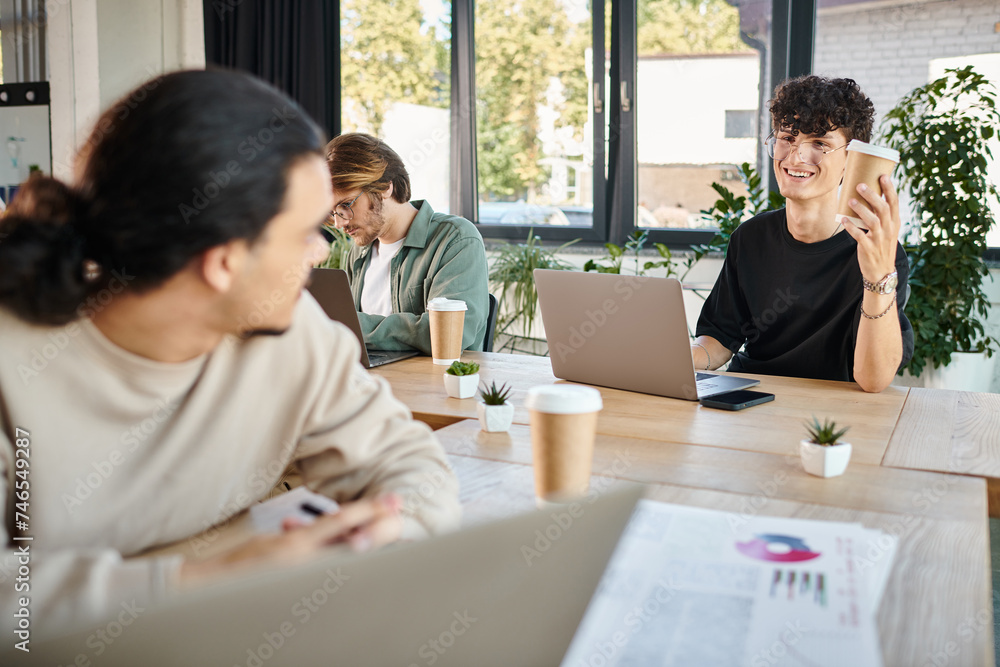 happy young men working on laptops in a coworking space, man in 20s offering coffee to colleague happy young men working on laptops in a coworking space, man in 20s offering coffee to colleague