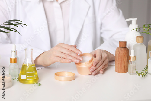 Dermatologist with jar testing cosmetic product at white table indoors, closeup