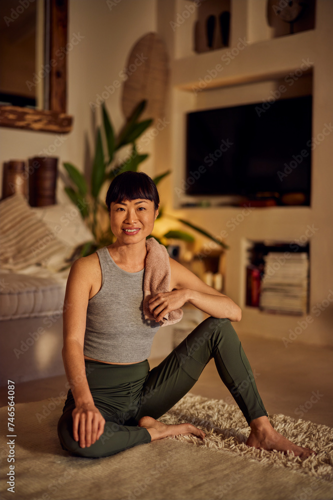 Fototapeta premium Portrait of a fit Asian woman, sitting on the floor, having a towel over her shoulder.