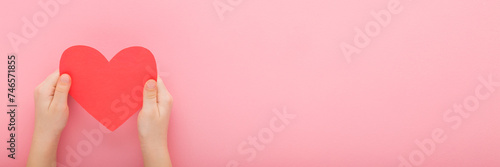 Little child hands holding red paper heart shape on pink table background. Pastel color. Love, health and hope concept. Closeup. Wide banner. Empty place for text. Point of view shot. Top down view.