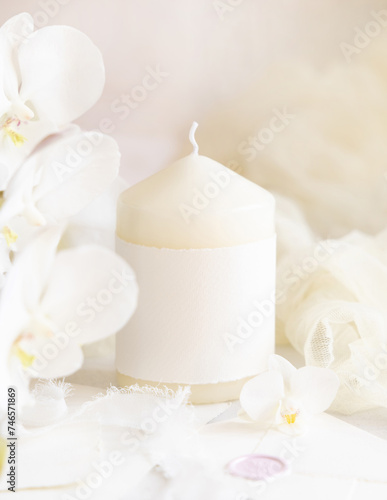 Pillar candle with a blank label near white orchid flowers close up, pastel romantic mockup