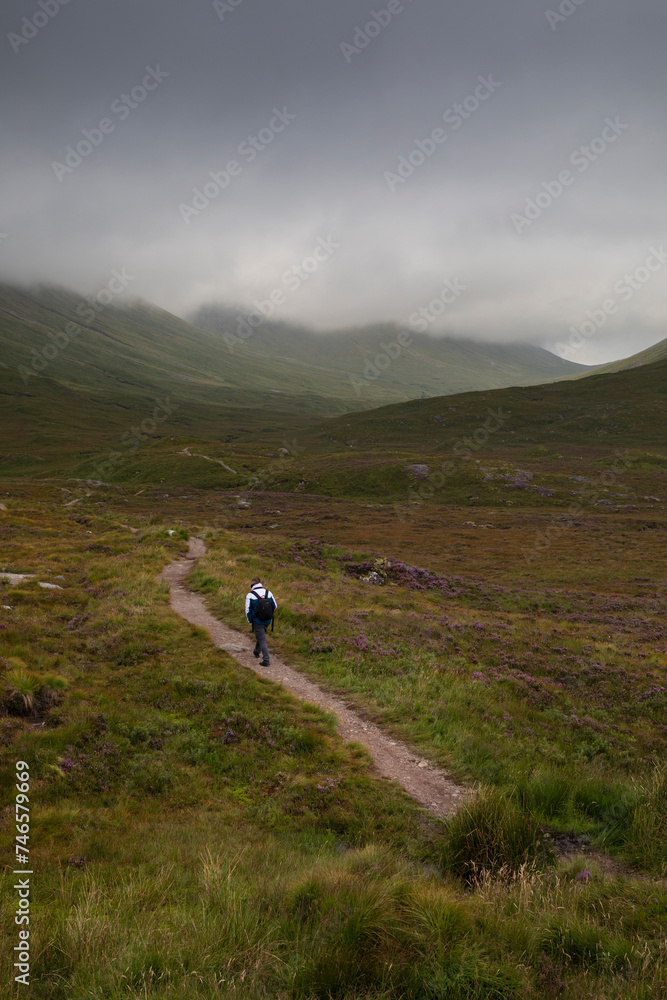 A man walks along a mountain path in the Glencoe Mountains on a day with dramatic skies from the Glencoe Lookout