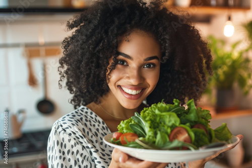Cute  young girl in kitchen holding plate with salad of fresh vegetables  herbs and smiling