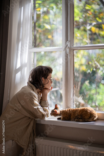 Lonely upset young woman leaning on windowsill near cat need psychologies help. Outcast by friends teen girl feeling frustration sadly looking at window loving cat trying emotional support to owner. photo