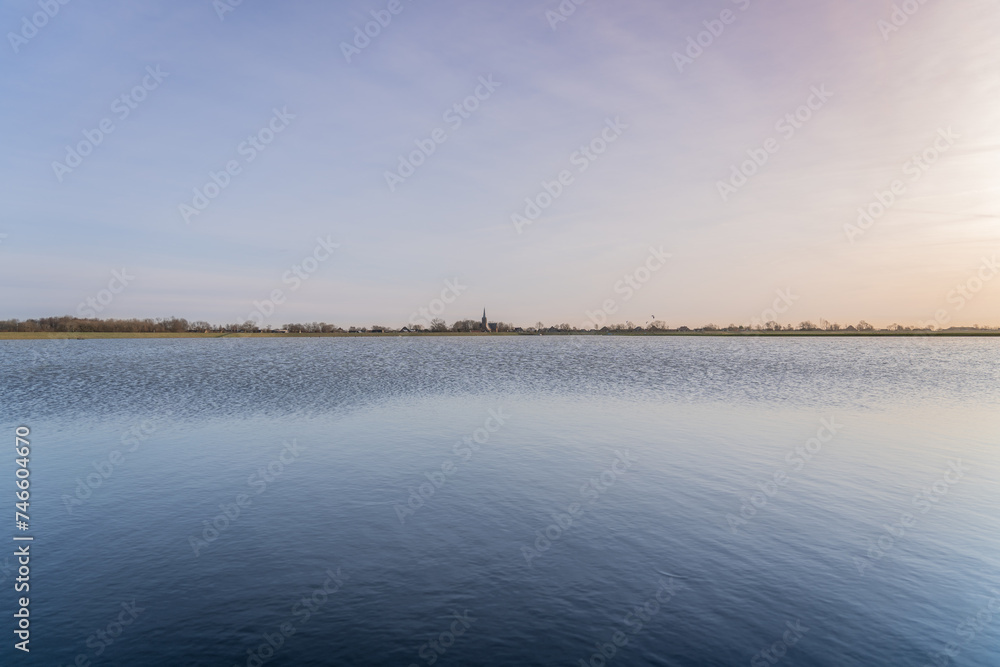Tranquil scene over a dutch village lake with overflowing water Into the meadow. Sky above transitions from a warm palette of sunset hues to the cooler tones reflecting on the placid waters surface.