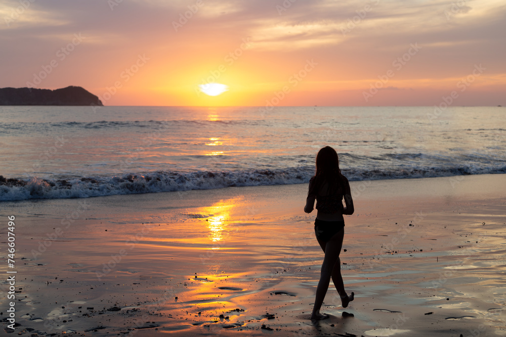 Silhouette back view of thin teenage girl walking on the beach along the edge of the Pacific Ocean during a fabulous pink sunset, Manuel Antonio, Costa Rica