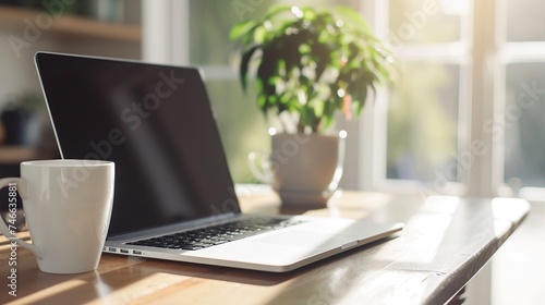 A white mug next to a laptop on a wooden table, illuminated by warm sunlight