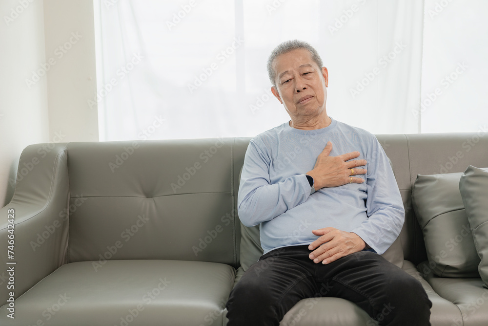 An old Asian man with chest pain was suffering from a heart attack, having difficulty breathing and clutching his chest from acute pain while sitting on the sofa at home. An old Asian man with chest pain was suffering from a heart attack, having difficulty breathing and clutching his chest from acute pain while sitting on the sofa at home.