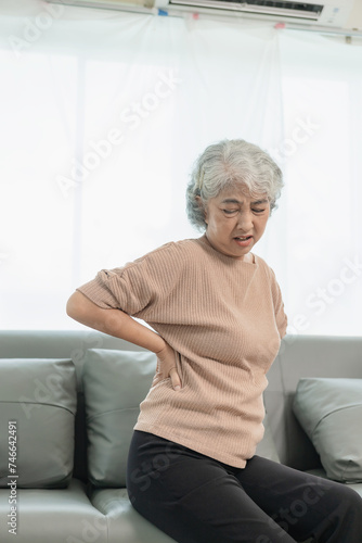 An elderly woman has back pain after waking up while sitting on the sofa at home. An elderly Asian woman has painful pain in her waist or buttocks after falling down.
