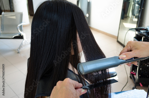 Planchado o alisado de cabello negro a clienta en Salón de Belleza con plancha eléctrica de pelo y peine. Servicio de peluquería profesional.  photo