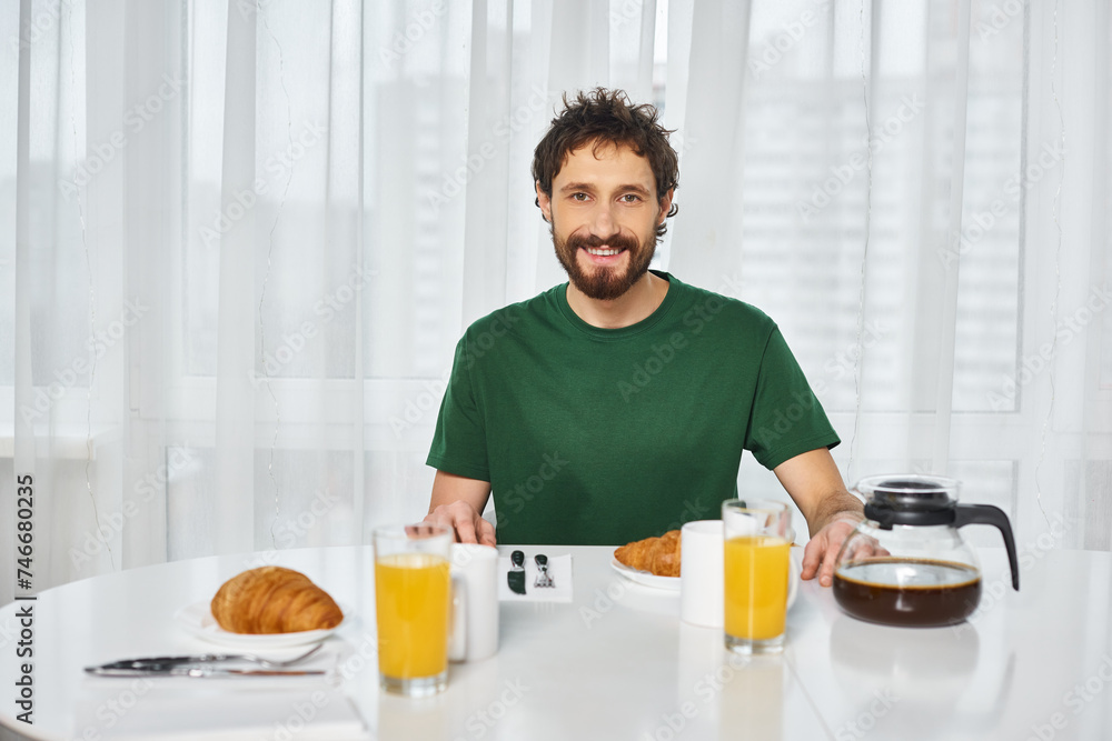 cheerful attractive man in casual attire sitting at table during breakfast and smiling at camera cheerful attractive man in casual attire sitting at table during breakfast and smiling at camera