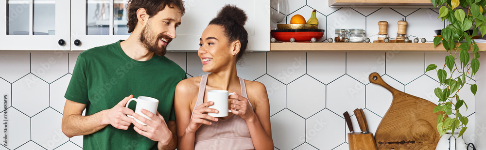 attractive jolly diverse couple in homewear enjoying hot coffee and looking at each other, banner attractive jolly diverse couple in homewear enjoying hot coffee and looking at each other, banner