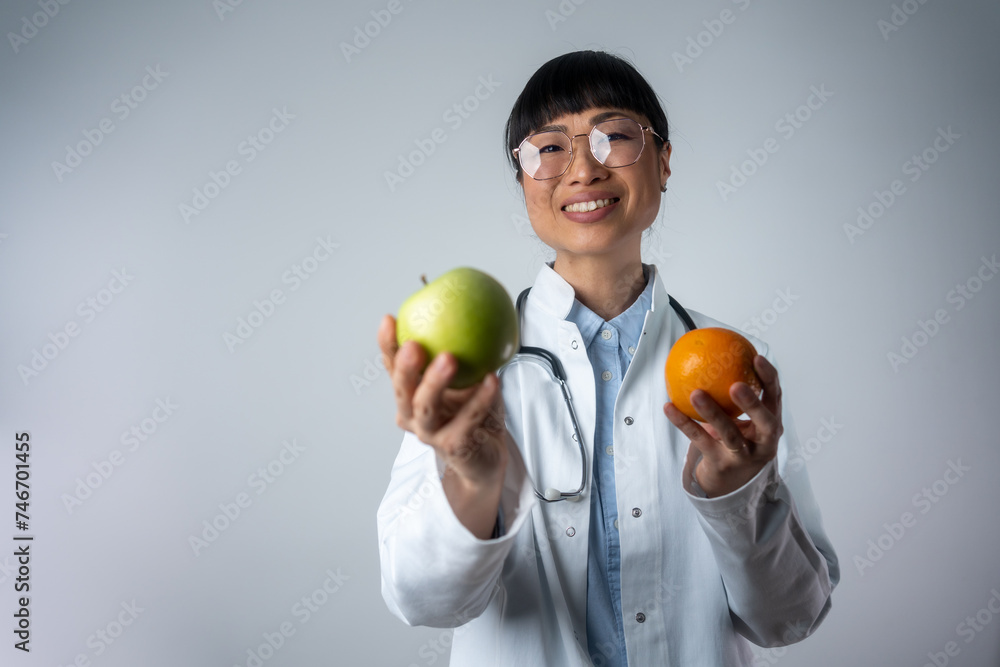 Smiling Asian dietician standing isolated and holding an orange and apple. Smiling Japanese woman doctor with a green apple and orange. Smiling Asian dietician standing isolated and holding an orange and apple. Smiling Japanese woman doctor with a green apple and orange.