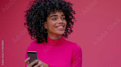 cheerful young woman with curly hair is holding a smartphone and looking away with a smile against a vibrant pink background.