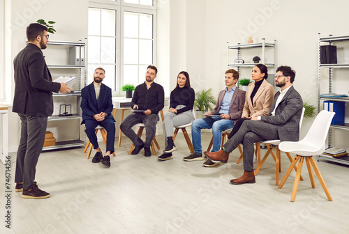 Smart confident male boss is having meeting with his subordinates at business meeting in office. Group of people sit on chairs in row and listen to leader about plan to improve business efficiency.