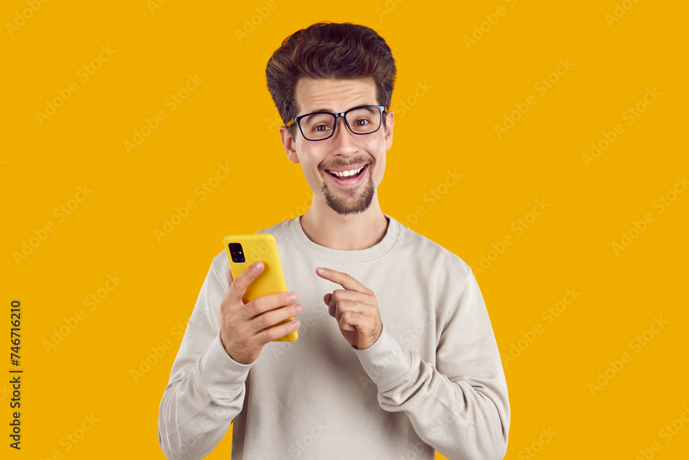 Portrait of young excited man pointing finger at smartphone. Cheerful guy in glasses and sweatshirt holding modern digital device and touching screen over yellow isolated studio background Portrait of young excited man pointing finger at smartphone. Cheerful guy in glasses and sweatshirt holding modern digital device and touching screen over yellow isolated studio background