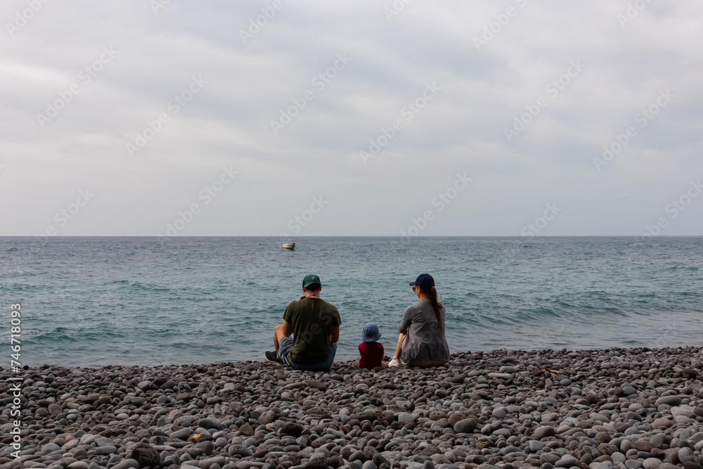 Family with small child sitting on idyllic volcanic black stone beach of Praia Garajai, Canico, Madeira island, Portugal, Europe. Waves hitting shoreline of majestic Atlantic Ocean. Vacation concept
