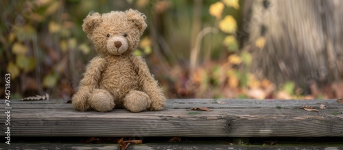 A close-up view of a brown teddy bear sitting comfortably on top of a sturdy wooden bench. The soft plush toy contrasts with the rough texture of the bench, creating a cozy and familiar scene.
