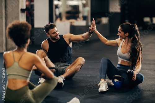 Group of three fit friends giving high five after exercise while sitting on a gym floor.