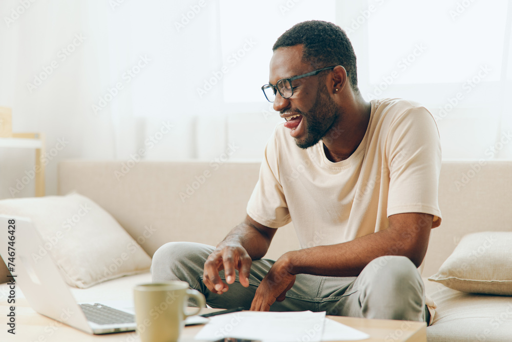 Smiling African American Man Working on Laptop at Home Sofa Happy young African American freelancer sitting on a modern couch in his home's living room He is typing on his laptop, fully immersed in