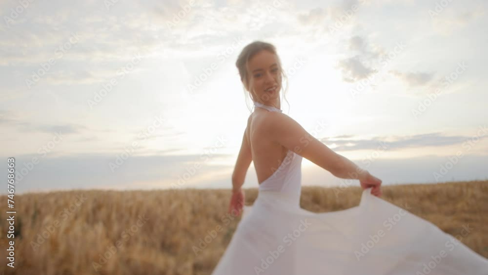 Elegant woman wearing flowing white dress billowing in wind moving amidst field of golden grass which stretching out into distance. Gently caucasian lady enjoying beautiful landscapes and sunset. Elegant woman wearing flowing white dress billowing in wind moving amidst field of golden grass which stretching out into distance. Gently caucasian lady enjoying beautiful landscapes and sunset.