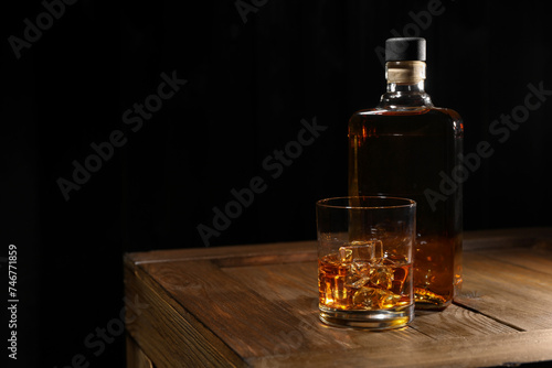 Whiskey with ice cubes in glass and bottle on wooden crate against black background, space for text © New Africa