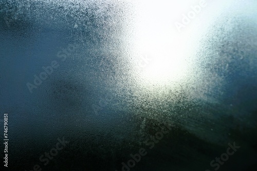Abstraktes Motiv mit beschlagener gefrorener Glasscheibe vor blaugrauem Himmel mit weißem Sonnenlicht und Haus bei Frost, Kälte und Sonne am Morgen im Winter