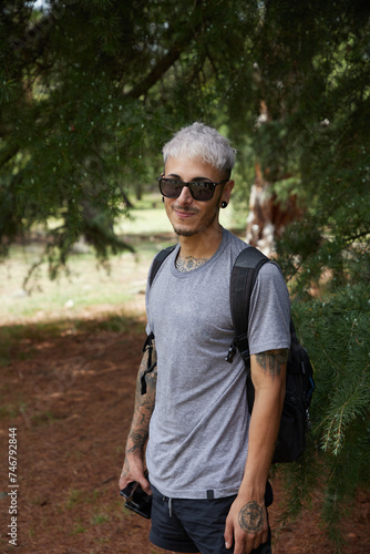 Hombre joven de pelo blanco con mochila mirando a camara plano americano en exterior fondo de bosque de pinos