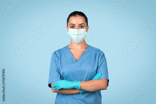 Young woman nurse in scrubs with mask and gloves on blue backdrop