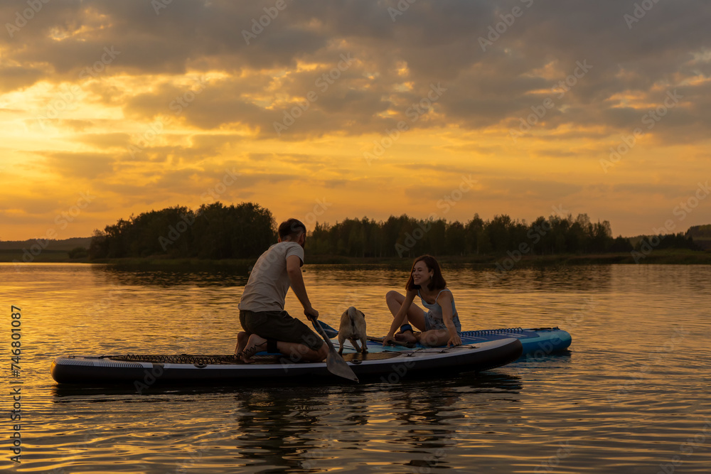 Happy couple paddle boarding at lake during sunset together with pug dog. Concept of active family tourism and supping with pets. Brave Dog Standing on SUP Board and enjoy lifestyle on summer vacation
