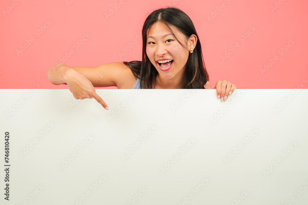Young asian woman pointing a blank empty board against red backdrop. Advice copy space mockup. Chinese teenage girl showing a down billboard copyspace at pink color background wall. Female presenting