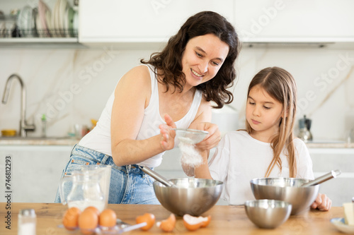 Mom and little girl prepare dough for baking, mom sifts and adds flour to bowl. Family activity, concept of happy family and parenting. Homemade food and little helper.