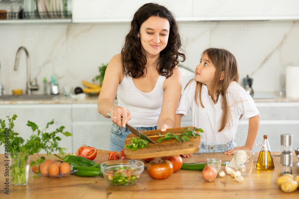 In kitchen, mom and daughter are cutting fresh vegetables to make salad. Groom explains to girl rules of cooking, peculiarities of using seasonings. Mother passing family recipe to her child.