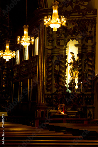 Interior de igreja católica com imagens de santos, e sequência de lustres acesos pendurados no teto. 