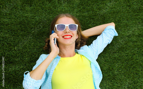 Happy smiling young woman calling on mobile phone while lying on grass in summer park © rohappy
