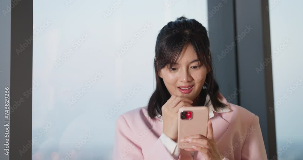 Joyful Asian woman with a charming smile engaging with her smartphone in a bright, modern indoor setting. Joyful Asian woman with a charming smile engaging with her smartphone in a bright, modern indoor setting.