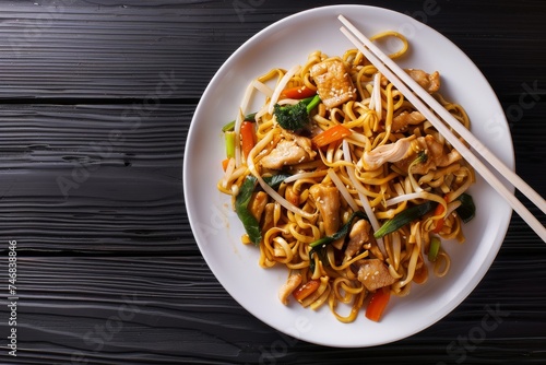 Chicken and vegetable udon stir fry on a white plate with chopsticks and sauce against a black wooden background