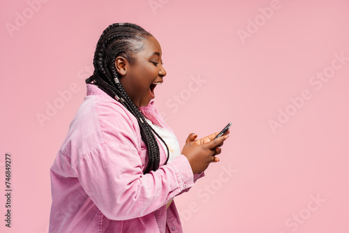 Happy African woman holding smartphone using mobile app, shopping online with sale, cash back isolated on pink background, copy space. Overjoyed female sports betting, win money, reading good news