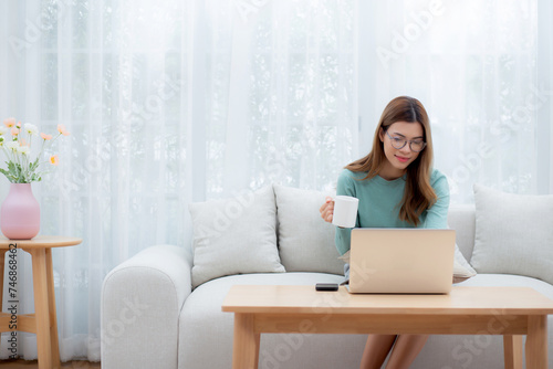 Young asian woman wearing glasses sitting sofa working on laptop computer and drinking coffee in living room at home, woman work from home with telework, freelance working, business and digital life. photo