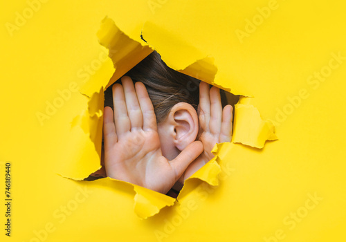 Female ear and hands close-up. Copy space. Torn paper, yellow background. The concept of eavesdropping, espionage, gossip and the yellow press.