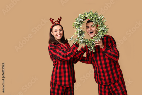Happy young couple in Christmas pajamas and with wreath on beige background