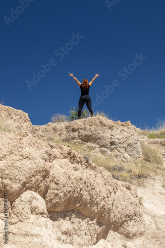 A person stands victorious atop a rugged summit, arms wide in jubilation against a clear blue sky, perfect for themes of success, freedom, and adventure.