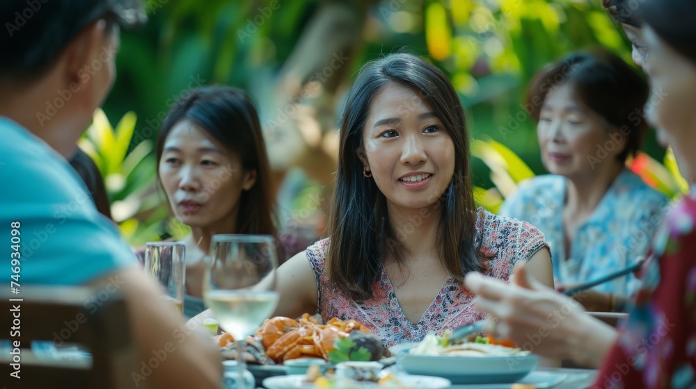 Fototapeta premium Young Asian women visit the family during parties outdoors in the garden. An attractive diverse group of people having dinner, eating food, and celebrating weekend reunion gathered at the dining table