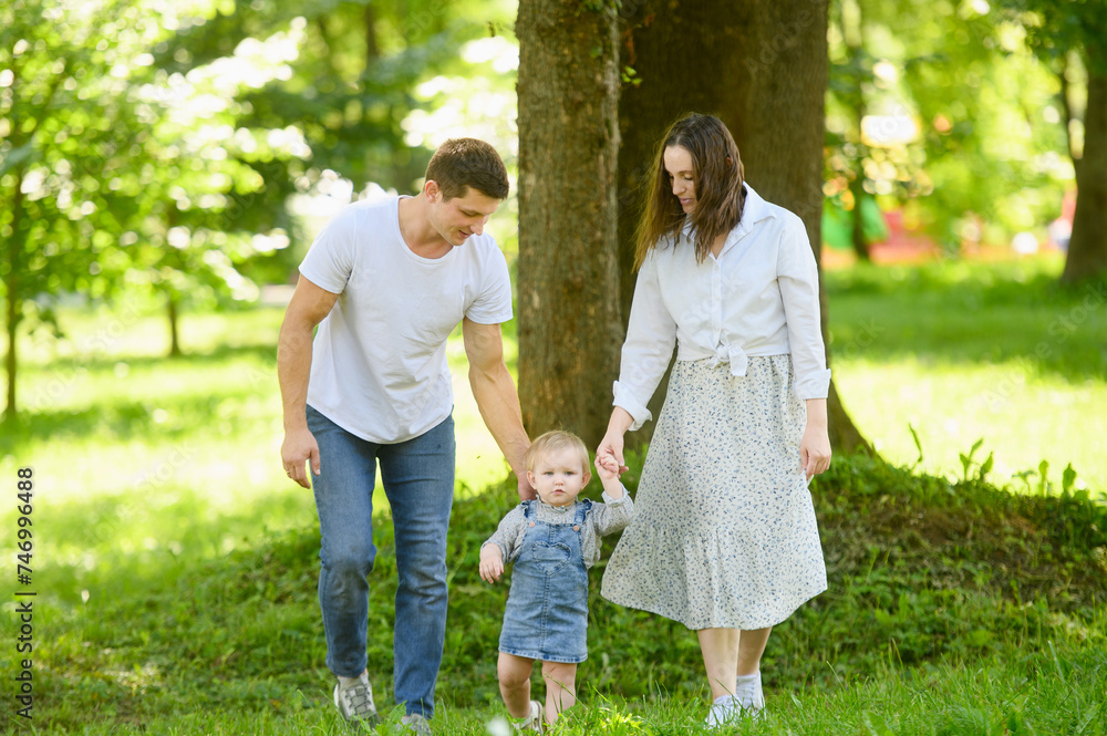 Child takes his first steps in the park on the grass, holding hands with his parents, learning to walk independently. Concept of family, family vacation in nature, first steps
